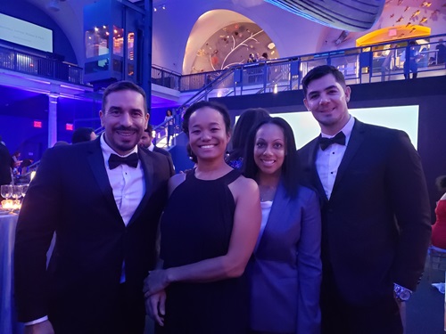 Four professionals posing for a photo at a gala with stairs, a balcony, and tables in the background
